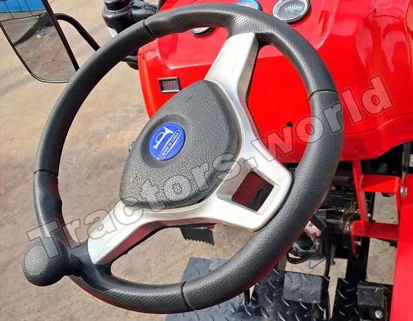Close-up of a red tractor cockpit: black steering wheel with a silver center and blue emblem, with dashboard gauges in the background.