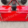 Red agricultural implement attached to a tractor, viewed from the front on a concrete surface with large tires visible above and a central hitch assembly.